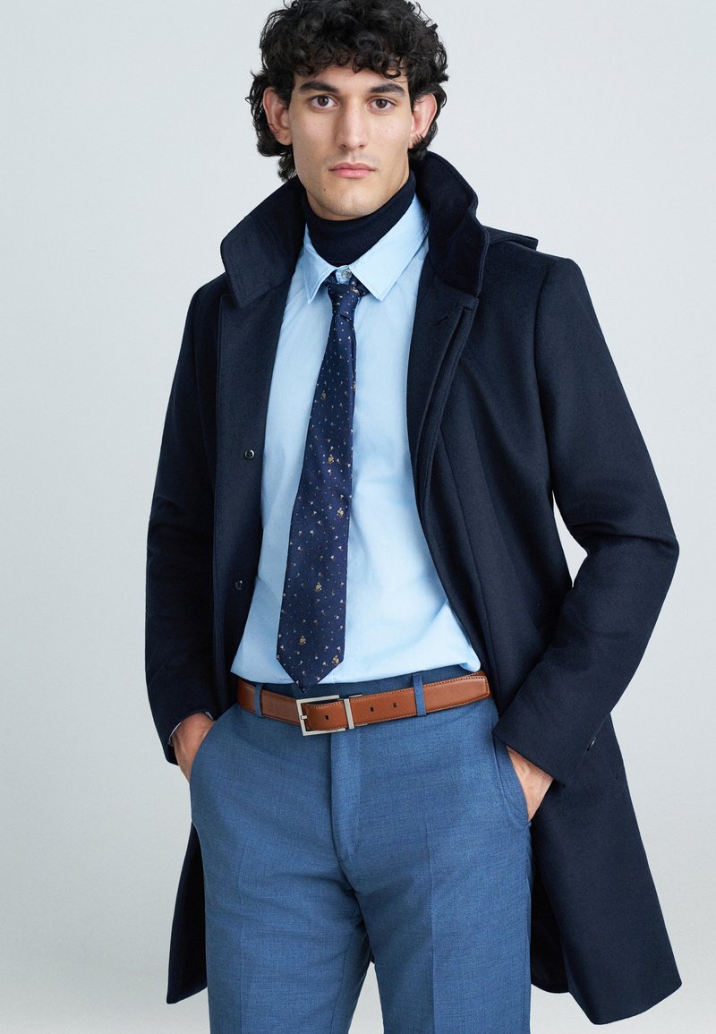 Cappotto in lana blu navy con colletto, abbinato a una camicia azzurro chiaro e una cravatta a fantasia. Cintura in pelle marrone con fibbia argentata, pantaloni blu.