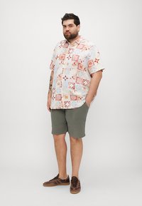 Chemise à manches courtes avec un design en patchwork multicolore, associée à un short vert olive et des baskets marron, mettant en valeur une coupe décontractée.
