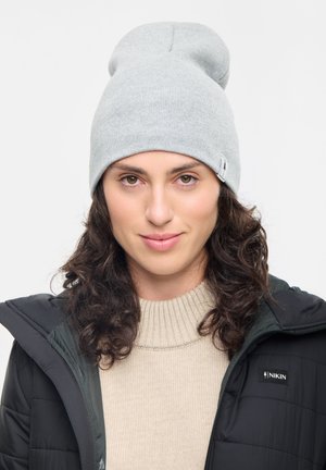 Jonge vrouw met krullend bruin haar die een lichtgrijze beanie, een beige trui en een zwart Nikin-jack draagt tegen een eenvoudige witte achtergrond.