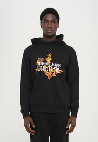 Črna kapuca s prominentnim natisom cvetja v oranžni in rumeni barvi ter napisom "VERSACE JEANS COUTURE". Mehak material, sproščen kroj.