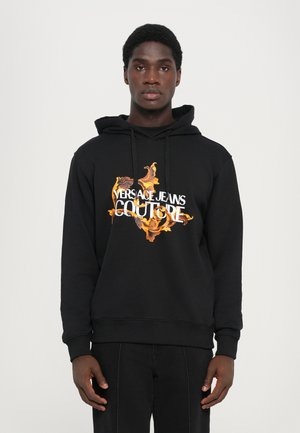 Mustaga kapuutsiga sviiter, mille esiküljel on silmapaistev oranž ja kollane lillemuster koos tekstiga "VERSACE JEANS COUTURE". Pehme materjal, mugav lõige.