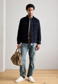 Navy jakna s gumbima preko karirane košulje, uparena s izblijedjelim plavim trapericama i bijelim tenisicama. Drži smeđu torbu sa zatvaračem i džepovima.