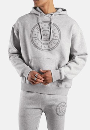 Mann trägt hellgrauen Reebok Classic Hoodie und passende Jogginghose mit Logos, Hände verschränkt, zeigt silberne Ringe an den Fingern.