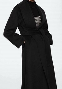 Manteau enveloppant en laine noire avec larges revers, manches longues et ceinture nouée à la taille, présentant une texture lisse et une coupe longue jusqu'au sol.