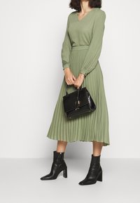 Abito plissettato verde oliva con maniche lunghe, scollatura a V, abbinato a una borsa strutturata nera e stivaletti. Tessuto morbido.