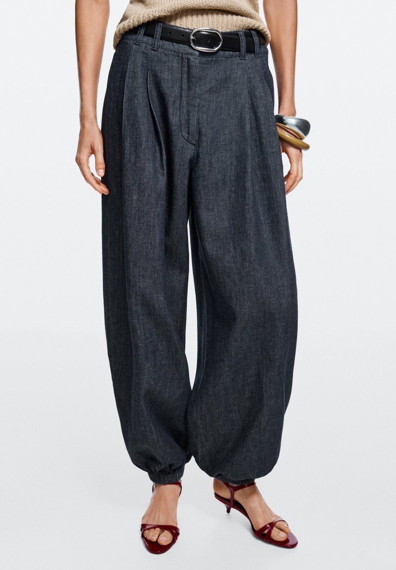 Wide-leg dark denim pants with elasticized cuffs, paired with a black belt, beige knit top, chunky bracelets, and red open-toe heels.