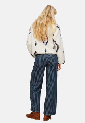 Veste en polaire crème avec motifs géométriques multicolores, associée à un pantalon ample à rayures bleues et des mocassins marron.