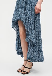 Πόδια που φορούν φούστα με λουλούδια μπλε χρώματος, υψηλής-χαμηλής γραμμής με βολάν και μαύρα ψηλοτάκουνα σανδάλια με λουράκια στον αστράγαλο, στέκονται σε λευκό φόντο.