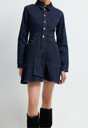 Kvinde iført mørkeblå denimkjole med lange ærmer, knapper foran, klaplomme og udvidet kant, kombineret med sorte knæhøje støvler.