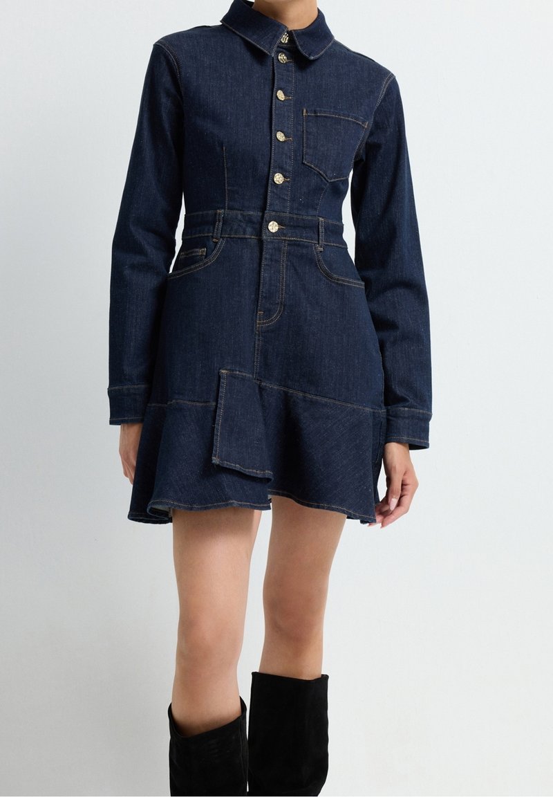 Femme portant une robe en jean bleu foncé à manches longues avec des boutons sur le devant, une poche plaquée et un ourlet évasé, associée à des bottes noires montantes jusqu'aux genoux.