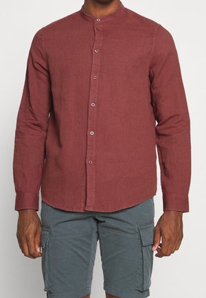 Man wearing a buttoned, long-sleeve, rust red mandarin collar shirt with gray cargo shorts against a plain background.