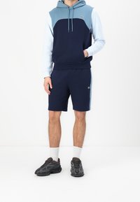 Mörkblå och ljusblå huvtröja med känguruficka, kombinerad med mörkblå shorts. Bär mörka sneakers och vita strumpor.