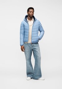 Man wearing light blue puffer jacket with hood, beige polo, wide-leg jeans, and white sneakers, standing with one hand in pocket.