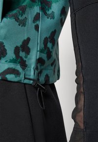 Haut en tissu à motif vert et noir avec un ourlet resserré, associé à un haut noir uni avec des détails transparents sur les manches.