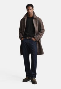 Brauner kariert Mantel, schwarzer Pullover mit Rundhalsausschnitt, dunkelblaue Jeans und braune Schnürschuhe. Der Mantel hat einen breiten Kragen und eine lockere Passform.