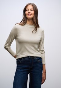 Béžový top s dlouhým rukávem a vysokým límcem, vyrobený z měkkého materiálu. Kombinovaný s tmavě modrými džíny s předními kapsami a klasickým střihem.