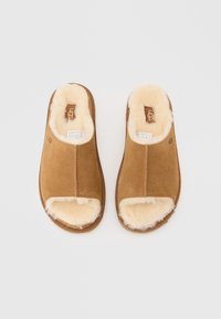 Paire de chaussons ouverts en daim beige avec doublure en shearling crème, semelles plates, et logo UGG sur la semelle intérieure et le côté.