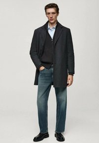 Grauem Mantel über einem dunklen Pullover, hellblauem Hemd und Jeans. Merkmale sind die gerade Passform und schwarze Lederschuhe. Saubere Linien und modernes Design.