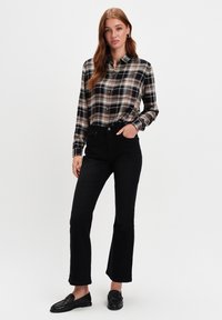 Młoda kobieta stoi z ręką w kieszeni, ubrana w beżową i czarną koszulę w kratę, czarne spodnie dzwony i czarne loafersy na białym tle.