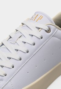 Λευκό δερμάτινο αθλητικό παπούτσι με μπεζ σόλα, λευκά κορδόνια και το λογότυπο "GAP" σε χρυσό στην γλώσσα.