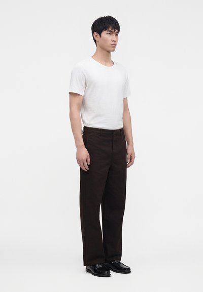 Jeune homme debout, portant un t-shirt blanc à manches courtes, un pantalon noir à jambes larges, et des chaussures habillées noires sur un fond blanc uni.