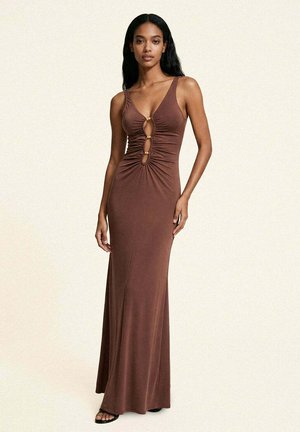 Donna che indossa un abito marrone senza maniche, lungo fino a terra, con tre fessure verticali e dettagli arricciati davanti, in piedi davanti a uno sfondo semplice.