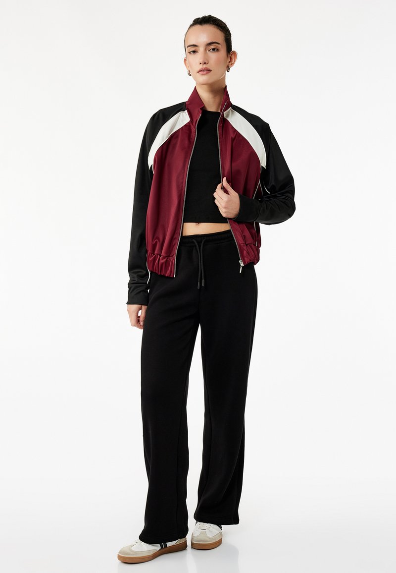Mörkröd och svart zip-up jacka med vita detaljer, tillsammans med svarta sweatpants. Har en figurnära midja och avslappnat snitt. Vita sneakers fulländar looken.