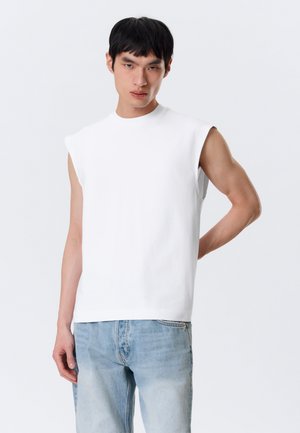 Junger Mann trägt ein weißes ärmelloses Oberteil und hellblaue Jeans, steht mit einer Hand hinter dem Rücken vor einem schlichten Hintergrund.