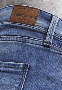 Blue denim jeans featuring a brown leather label with "Pepe Jeans" embossed. The fabric has a slightly faded texture with stitched accents.
