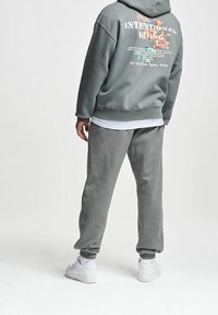 Personne portant un sweat à capuche gris avec un motif floral et du texte dans le dos, un pantalon de jogging gris, des baskets blanches, debout sur un fond blanc.