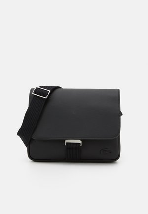 Zwarte, getextureerde crossbodytas met verstelbare brede band en zilveren gesp, met een klein reliëf krokodillenlogo aan de voorkant.