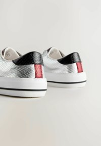 Para srebrnych sneakersów z czarnymi teksturowanymi wkładkami na pięcie i czerwonymi pionowymi paskami na białych podeszwach z czarnym wykończeniem, pokazanych z tyłu na białej powierzchni.