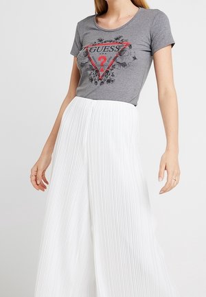 Femme portant un t-shirt logo Guess gris avec un design floral et un pantalon plissé large taille haute blanc, sur fond uni.