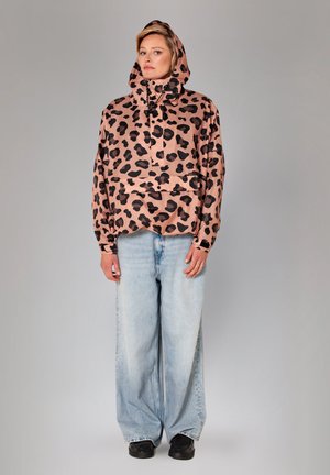 Vrouw draagt een roze-zwart luipaardprint capuchonjas, lichtblauwe wijde jeans en zwarte schoenen, staand tegen een effen grijze achtergrond.