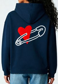 Felpa blu navy con un grande cuore rosso e un fermaglio di sicurezza bianco ricamati sul retro. Presenta una tasca a marsupio e polsini a coste.