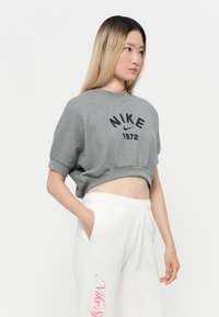 Harmaa lyhythihainen huppari, jossa on mustalla painettu "NIKE 1972", yhdistetty valkoisiin collegehousuihin, joissa on vaaleanpunainen teksti. Pehmeä puuvillasekoitekangas.