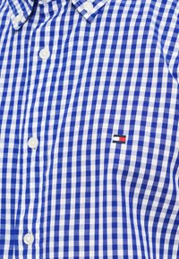 Μπλε και λευκό καρό πουκάμισο από ύφασμα, με λευκά κουμπιά και μικρό λογότυπο με κόκκινες, λευκές και μπλε λεπτομέρειες.