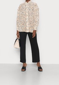 Blouse florale à manches longues avec épaules à volants en crème, pantalon large noir, et sandales à glissière noires ; tenant un sac à main bicolore.