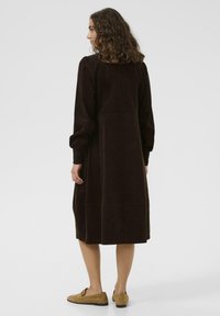 Braunes Cordkleid mit langen Ärmeln, knielangem Saum und einem taillierten Oberteil. Strukturierter Stoff mit dezenten Nähten. Kombiniert mit hellbraunen Schuhen.