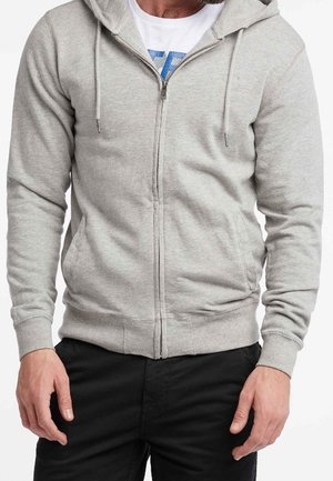 Gray zip-up hoodie in soft material with a drawstring hood, ribbed cuffs and hem, and front pockets. Worn over a white T-shirt.