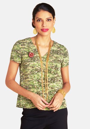 Grün gemusterte Bluse mit V-Ausschnitt, kurzen Ärmeln und einer Schmuckbrosche als Akzent. Kombiniert mit einem schwarzen Unterteil und goldenen Accessoires.