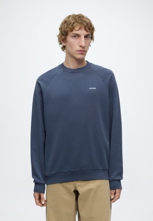 Homme blond portant un sweat-shirt bleu marine à manches longues avec le texte « LES DEUX » sur la poitrine et un pantalon beige, debout sur fond blanc.