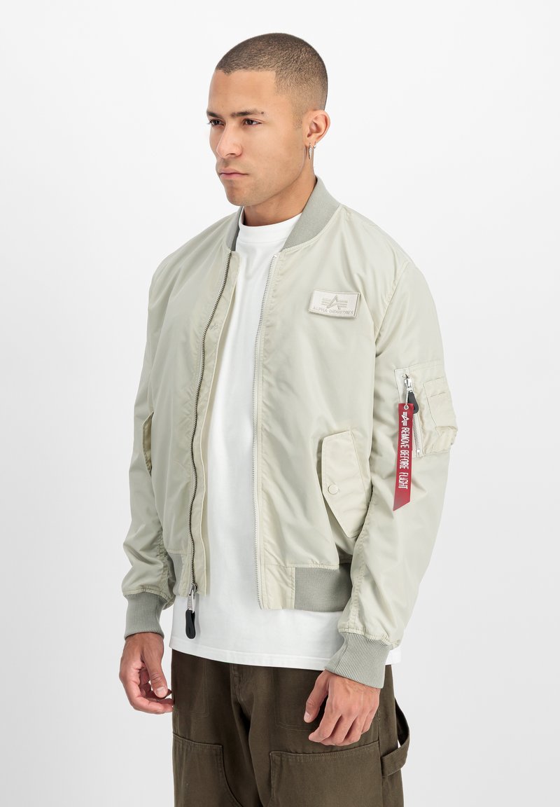 Blouson aviateur gris clair avec fermeture éclair à l'avant, poignets côtelés et poches. Comprend une étiquette rouge sur la manche gauche et un patch logo en tissu sur la poitrine.
