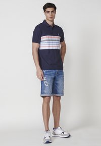 Jūrinės spalvos polo marškiniai su horizontaliomis juostelėmis raudonos, baltos ir mėlynos spalvos, derinami su nusidėvėjusiais džinsiniais šortais ir baltais sportiniais bateliais.