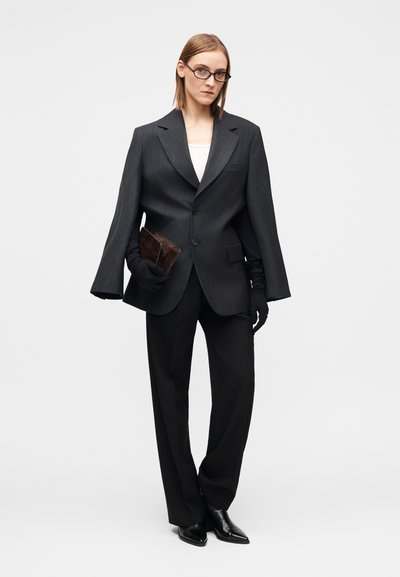 Overdimensioneret mørkegrå blazer med tynde striber og brede ærmer, kombineret med sorte bukser og sorte spidse hæle. Holder en brun clutchtaske.