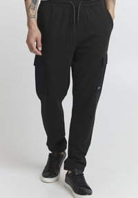 Black sweatpants with a drawstring waist, featuring side cargo pockets in a contrasting dark material and ribbed ankle cuffs.