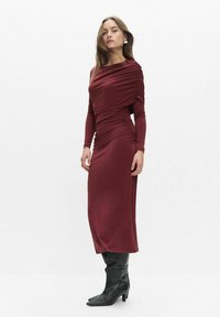 Bordowa sukienka z długim rękawem, z drapowaniami i dopasowanym krojem, wykonana z gładkiego materiału, zestawiona z czarnymi kozakami sięgającymi kolan.