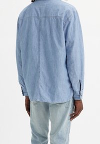 Chemise bleu clair à manches longues en denim avec une finition légèrement texturée, dotée de poignets à boutons et d'un ourlet courbé. Vue de dos.
