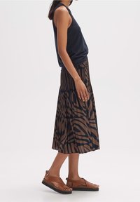 Kvinna klädd i en ärmlös marinblå top, brun och svart mönstrad veckad midikjol samt bruna lädersandaler med svarta sulor.