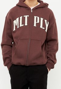 Felpa con zip color bordeaux, con testo ricamato bianco "MLT PLY", tasche a canguro e tessuto testurizzato. Presenta un cappuccio e polsini a costine.
